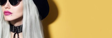 Güneş gözlüğü takan, siyah şapkalı ve boyunluk takan genç sarışın kızın stüdyo portresi. Kopyalama alanı olan sarı arkaplan.