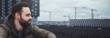 Şehir binalarının arka planında çatıda oturan sakallı genç adamın yakın plan portresi. Boşluk konseptini kopyala.