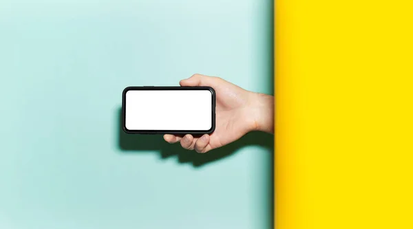 Close-up of male hand holding horizontal smartphone with mockup between two studio backgrounds of yellow and aqua menthe colors.