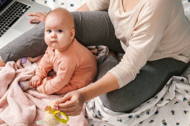 Dizüstü bilgisayarı olan bir anne ve bebek yatakta çalışıyor. Anne karantina sırasında evde çocukla birlikte çalışıyor. Bebek oynarken tanınmayan bir annenin yatakta oturup laptopta çalışmasına yakın plan.