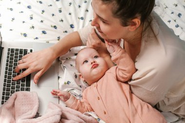 Küçük bebeğiyle yatakta oynaşan genç bir anne. Evde dizüstü bilgisayarı ve yeni doğmuş bebeğiyle genç bir anne. Çocuklar ve aletler. Ev rahatlığı. Dikkat ve dikkat. Evden çalışıyorum. Kadın işi..