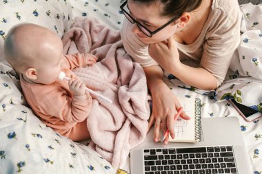 Küçük bebeğiyle yatakta oynaşan genç bir anne. Evde dizüstü bilgisayarı ve yeni doğmuş bebeğiyle genç bir anne. Çocuklar ve aletler. Ev rahatlığı. Dikkat ve dikkat. Evden çalışıyorum. Kadın işi..