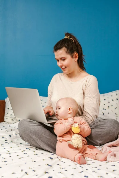 Young mother with her little baby relaxing and playing in the bed. Young mother in home office with laptop and her newborn baby. Children and gadgets. Home comfort. Care and attention. Work from home. Female business.