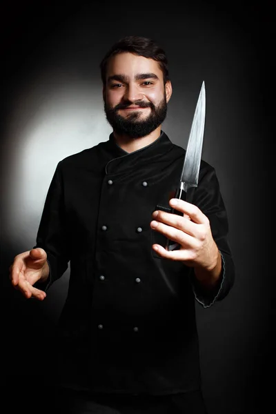 Chef professionista con la barba in uniforme nera con un coltello da cucina in mano su sfondo nero — Foto Stock