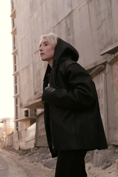 Une fille blonde avec une coupe de cheveux courte, en vêtements noirs à la mode et un sac se promène dans la rue — Photo