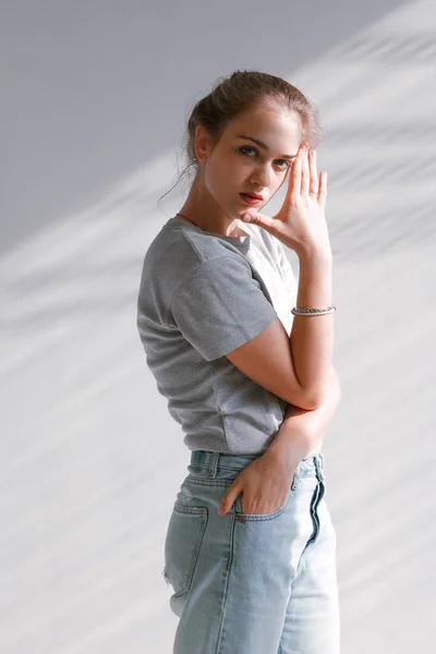 A beautiful, sweet girl in jeans and a sweatshirt stands near a white wall — Stock Photo, Image
