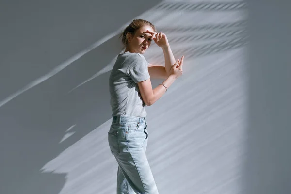 Uma menina bonita e doce em jeans e uma camisola fica perto de uma parede branca — Fotografia de Stock