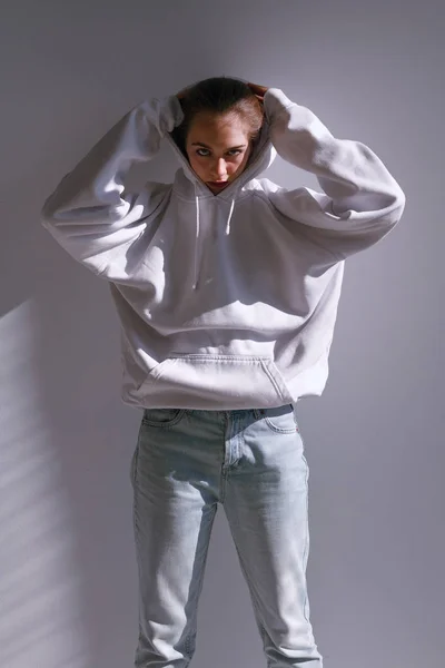 Uma menina bonita e doce em jeans e uma camisola fica perto de uma parede branca — Fotografia de Stock
