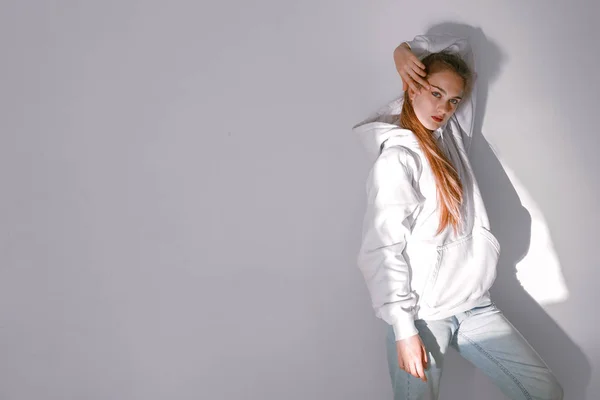 Uma menina bonita e doce em jeans e uma camisola fica perto de uma parede branca — Fotografia de Stock