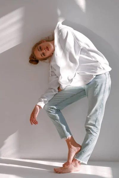 Una hermosa y dulce chica en jeans y una sudadera se encuentra cerca de una pared blanca — Foto de Stock