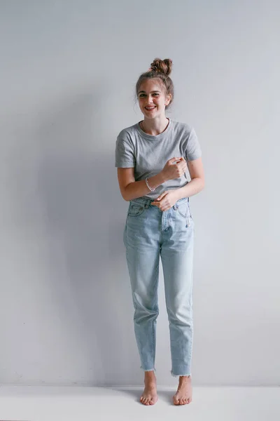 Uma menina bonita e doce em jeans e uma camisola fica perto de uma parede branca — Fotografia de Stock