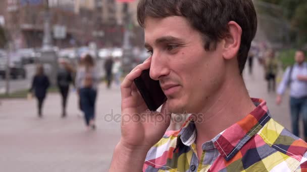 Χαμογελαστός νεαρός άνδρας μιλά για ένα κινητό τηλέφωνο. Φόντο πόλη, ανθρώπους, αυτοκίνητα. Ευρώπη — Αρχείο Βίντεο