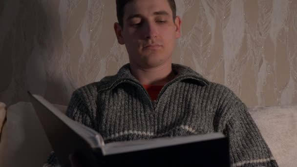 英俊的男人坐在沙发上读圣经书 晚上的房间 慢动作 — 图库视频影像