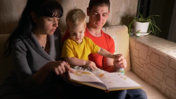 幸福的家庭 年轻男子和妇女在沙发上看书与孩子 慢动作 晚上房间 — 图库视频影像