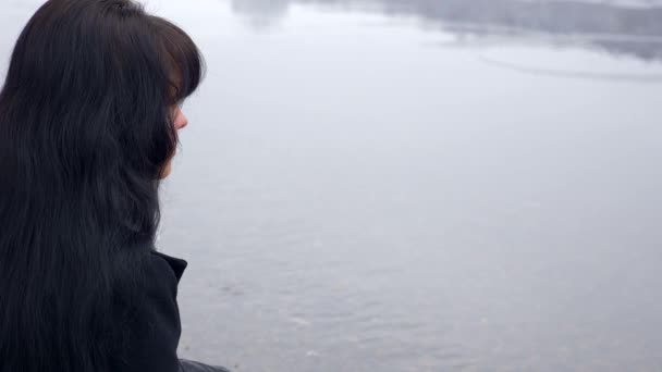 Krásná bruneta sedí na břehu řeky myšlení a pohledu ve vodě. Zpomalený pohyb zimní