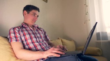 Laptop sörf Internet kullanan genç adam kanepe kanepeye oturur. Ağır çekim 30p 0,5 gerçek zamanlı hız 60p