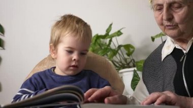 Büyük büyükannem torununun torunuyla kitap okuyor. Aktif Yaşlı Kadın, Küçük Çocuk 'la Bebek Bakıcılığı Evde Gündüz Vaktinde Bir Kitabın Sayfalarında Resimlerini Gösteriyor. Yavaş çekim 0. 5 hız 60 fps