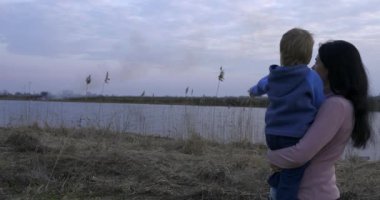 Anne ve Oğlu Kuru Çimen Ovası 'nda Yaban Ateşi' ni izliyor. Nehrin yanındaki arazide yürüyüş ve yürüyüş. Kırsalda Sabah Akşamı. Kırsal Manzara Manzarası Bahar Sezonu