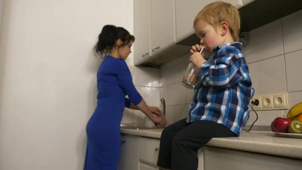 Kind Trinkt Fruchtcocktail Sohn Sitzt Auf Küchenarbeitsplatte Mutter Wäscht Glas — Stockvideo