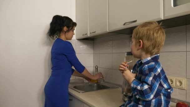 Kind Trinkt Fruchtcocktail Sohn Sitzt Auf Küchenarbeitsplatte Mutter Wäscht Glas — Stockvideo