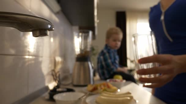 年轻的母亲用水给儿子斟满杯子 小孩坐在厨房台面上 晨光透过窗户照进来 2X慢动作 5速度60 Fps — 图库视频影像