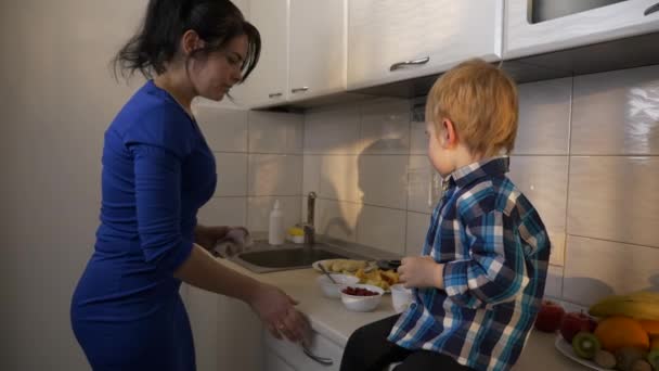 息子のための高速朝食を準備忙しい若い母親 キッチンカウンターの上に座っている子供 窓から朝日が差し込む朝 2倍スローモーション 5速度60 Fps — ストック動画