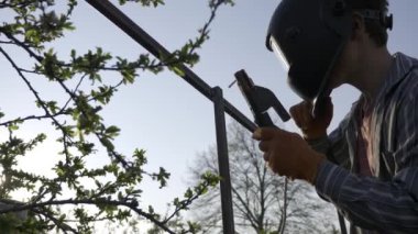 Arka bahçede Genç Kaynak Metal İnşaat. Koruyucu Miğferde çalışan bir işçi. Kırsal kesimdeki kır evinin yanındaki bahçe. 4K 10-bit Hibrit Günlük Gama HLG HDR