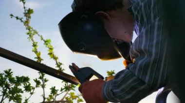 Arka bahçede Genç Kaynak Metal İnşaat. Koruyucu Miğferde çalışan bir işçi. Kırsal kesimdeki kır evinin yanındaki bahçe. 4K 2x Yavaş çekim 0. 5 x 60 FPS 