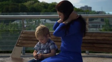 Anne, çocuğuyla birlikte şehir parkındaki Bench 'te oturuyor. Şehir Yeşil Alanı. Parlak Güneşli Yaz Günü