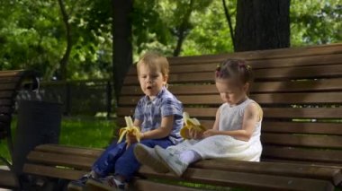Mutlu Çocuk ve Kız Park 'ta Bench' te oturuyor. Çocuklar muz yer. Parlak Güneşli Yaz Günü