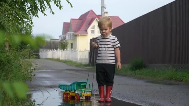 Little Boy Walking and Playing with Toy Truck Vehicle in Water Puddle After Rain. Evening Suburbs. 2x Slow motion - Half Speed 60 FPS — Stock Footage Little Boy Walking Playing Toy Truck Vehicle Water Puddle Rain Royalty Free Stock Video