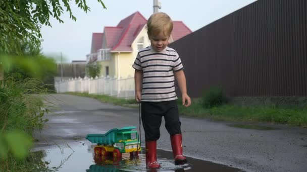 Little Boy Walking and Playing with Toy Truck Vehicle in Water Puddle After Rain. Evening Suburbs. 2x Slow motion - Half Speed 60 FPS — Stock Footage Little Boy Walking Playing Toy Truck Vehicle Water Puddle Rain Royalty Free Stock Video