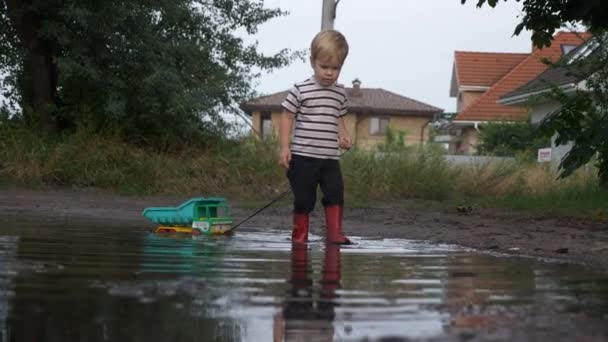 Little Boy Walking and Playing with Toy Truck Vehicle in Water Puddle After Rain. Evening Suburbs. 2x Slow motion - Half Speed 60 FPS — Stock Footage Little Boy Walking Playing Toy Truck Vehicle Water Puddle Rain Royalty Free Stock Footage