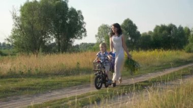 Küçük Çocuk Kırsal Yol 'da Dolaşan Genç Anne' ye yardım ederek Çocuk Bisikleti Sürmeyi Öğreniyor. Yaz Akşamı. 2x Slow motion - Yarı hız 60 FPS