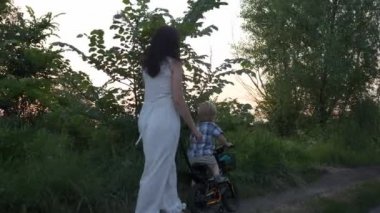 Küçük Çocuk Kırsal Yol 'da Dolaşan Genç Anne' ye yardım ederek Çocuk Bisikleti Sürmeyi Öğreniyor. Yaz Akşamı. 2x Slow motion - Yarı hız 60 FPS