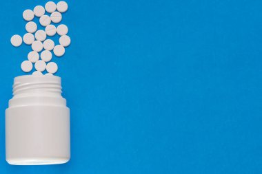 Beyaz tabletler mavi arka plandaki bir şişeden dağılıyor.