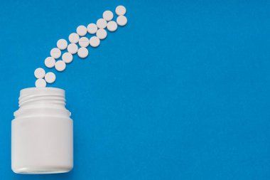 Beyaz tabletler mavi arka plandaki bir şişeden dağılıyor.
