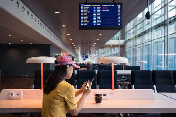 从侧面看亚洲女人在机场的帽子冲浪手机和用一次性帽子喝咖啡 — 图库照片