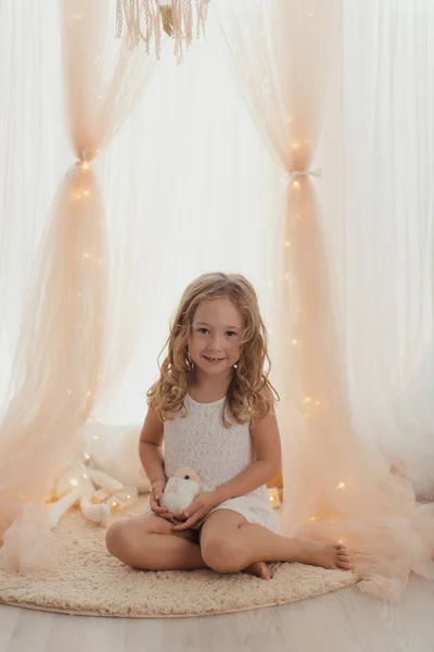 温柔的小女孩 身穿白衣 坐在毛毯上 头戴玩具 面带微笑 在童话般的灯光和时髦的窗帘前看着镜头 — 图库照片