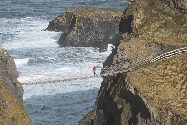 Tourist walking on rope bridge suspended between cliffs in Northern Ireland — Stock Photo, Image