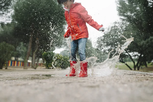 在灰蒙蒙的日子里 穿着红色雨衣和橡胶靴的可爱的快乐的孩子在公园的水坑里跳来跳去 — 图库照片