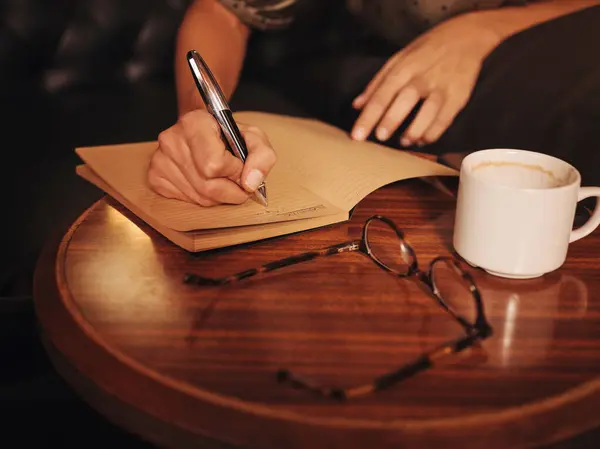 上記の作物から認識できない女性は カフェでコーヒーとガラスと丸い木製のテーブルでノートブックにペンで書く — ストック写真