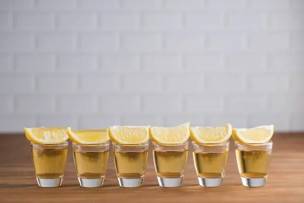金色のテキーラとぼやけた背景に白い壁と木製のテーブルの上にレモンのスライスを持つガラスのスポットの行 — ストック写真