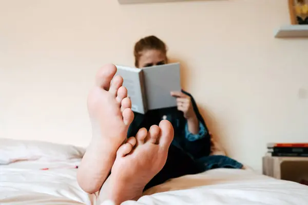 認識できない裸足の女性でカジュアル服読書本で関心を持ちながらリラックスしながら一人で柔らかいベッドの上に安全に家にいます — ストック写真
