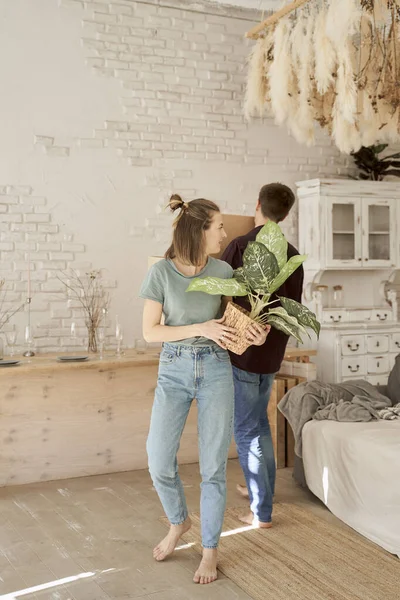 Босонога жінка, що носить горщик з квіткою і дивиться на чоловіка в кімнаті, де меблює новий будинок — стокове фото