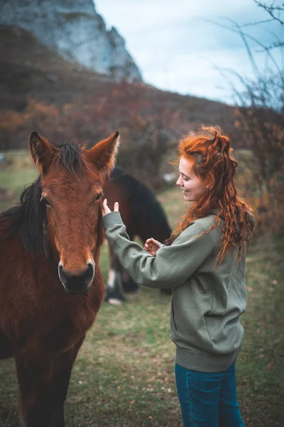 Vue latérale d'une femme souriante aux longs cheveux roux à capuche caressant un cheval brun avec crinière noire dans un pâturage de montagne — Photo de stock