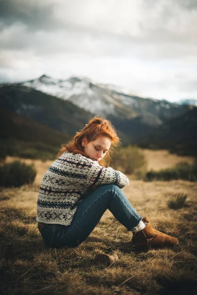 Seitenansicht einer nachdenklichen, charmanten Frau mit roten Haaren im warmen Pullover, die bei grauen Tagen mit geschlossenen Augen auf dem Rasen in den Bergen sitzt — Stockfoto