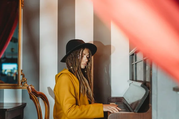 Vista laterale di una donna hipster seria con dreadlocks che indossa un cappotto giallo e un cappello nero che suona il pianoforte mentre è seduta in una stanza in stile retrò — Foto stock