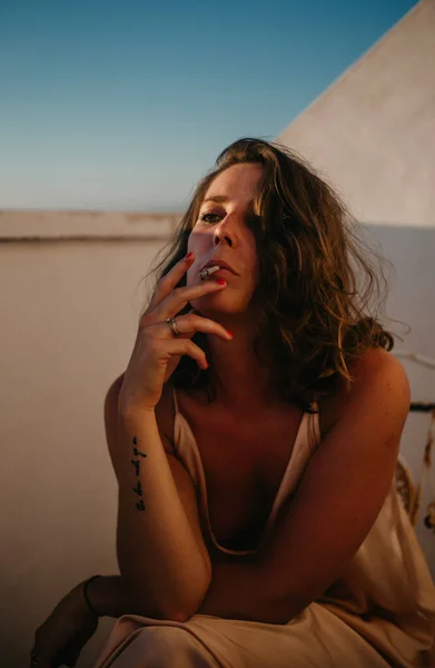Friedliche Frau sitzt am Zaun des Balkons und raucht Zigarette mit Sonnenuntergang auf verschwommenem Hintergrund — Stockfoto