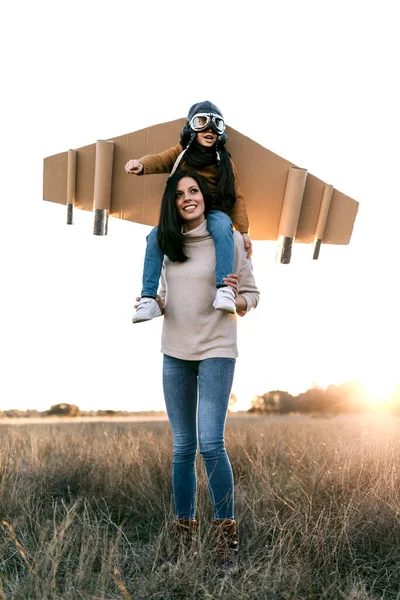 Happy cute boy wearing goggles and cardboard wings while sitting on mother shoulders and imitating aviator on meadow in backlit — Stock Photo